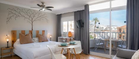 A hotel room with a bed, a dining table with chairs, a TV, and a balcony with a view of the city.