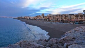 A coastal beach with a rocky foreground, a sandy beach, and a row of buildings in the background. - Palm Beach Club (Torremolinos)