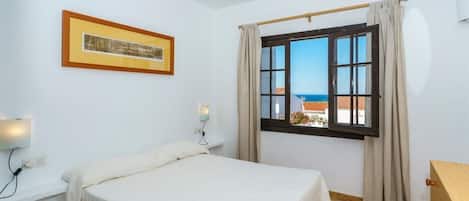 Un dormitorio con cama, ventana con cortinas, lámpara de pared e imagen enmarco.