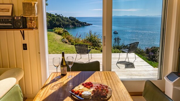 Une salle à manger avec une table dressée pour deux personnes, une bouteille de vin et une assiette de fromage et de raisins. Dehors, on peut admirer un paysage magnifique avec une côte et un bateau sur l’eau.