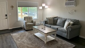 A living room with a grey sofa, a patterned armchair, a wooden coffee table, and a large window with blinds.