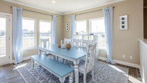 A dining area with a blue table and white chairs, a patterned rug, and large windows with curtains.