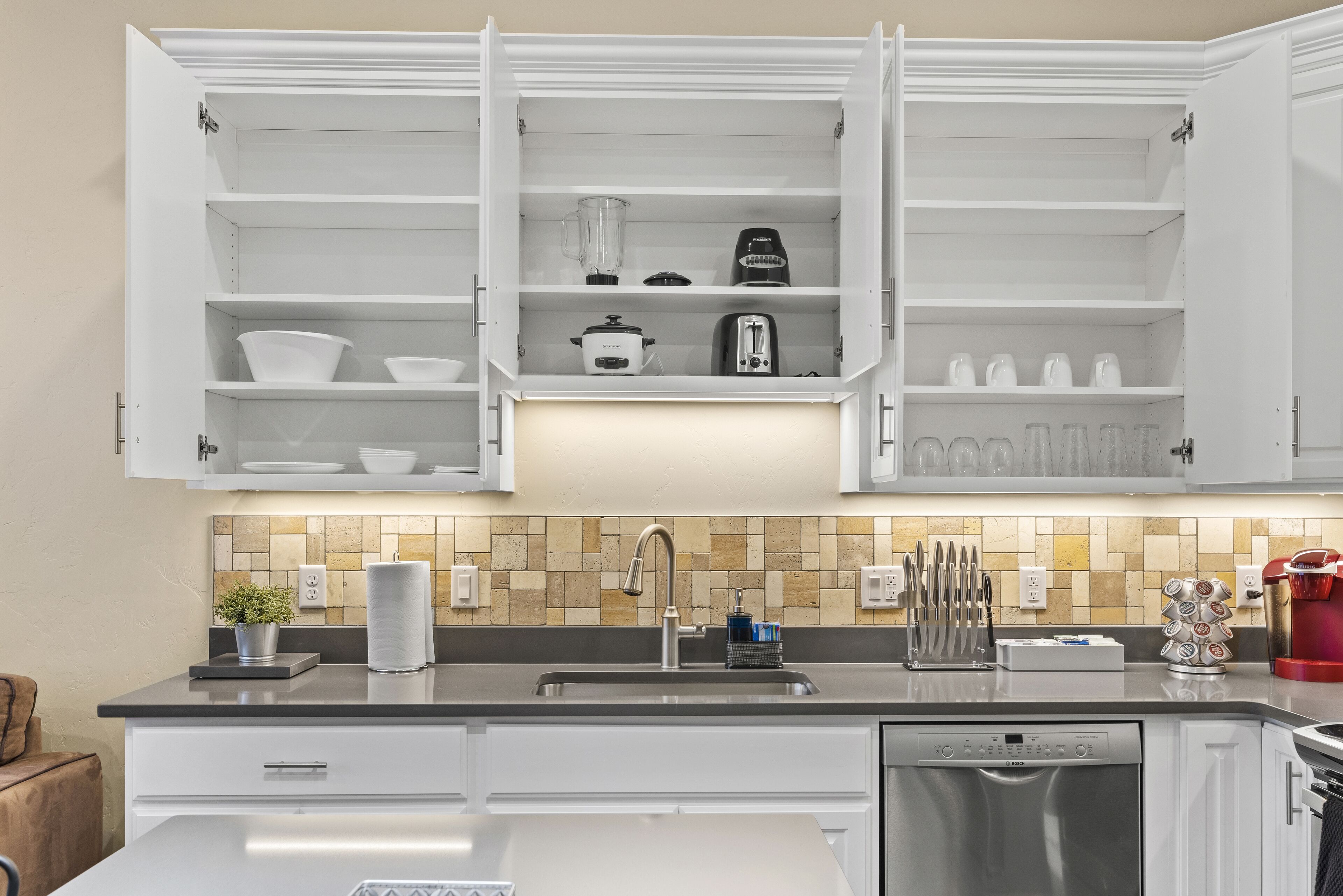 Uma cozinha moderna com armários brancos, painel de azulejos e lava-louças de aço inoxidável.
