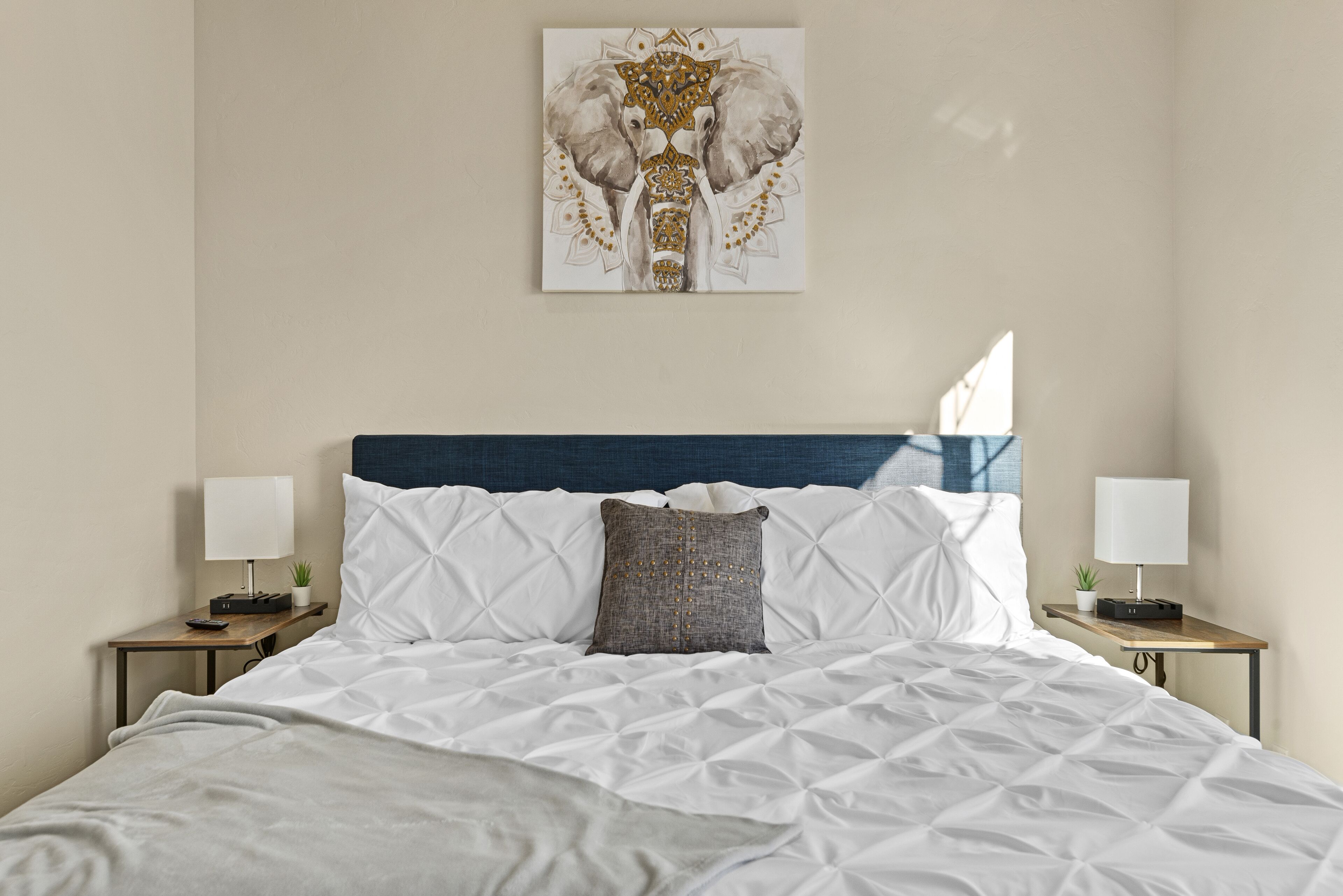 Un lit bien fait, avec une couette blanche, une tête de lit bleue et une œuvre d’art représentant un éléphant au-dessus. Deux tables de chevet, chacune agrémentée d’une lampe et d’une petite plante, se trouvent de part et d’autre du lit.