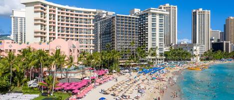 A beachfront hotel with multiple high-rise buildings, a sandy beach with umbrellas and lounge chairs, and palm trees.