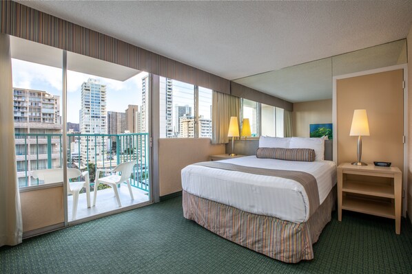 Habitación de hotel con una cama grande, un balcón con sillas y vistas a edificios de la ciudad.