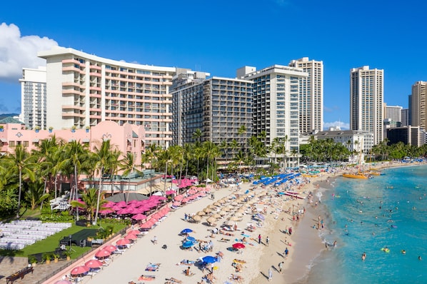 A beachfront hotel with multiple high-rise buildings, a sandy beach with umbrellas and lounge chairs, and palm trees.