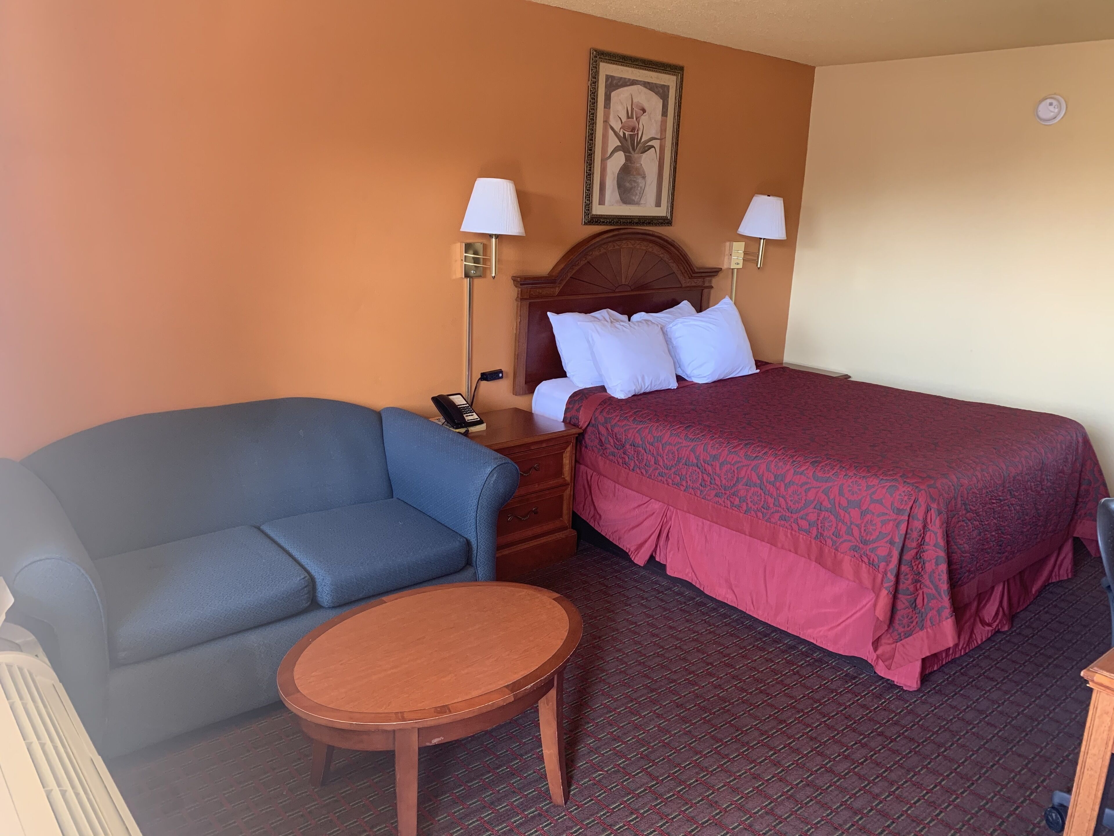 Une chambre d’hôtel avec un canapé bleu, une table en bois, un lit recouvert d’une couverture rouge, deux lampes de chevet et un tableau encadré au mur.
