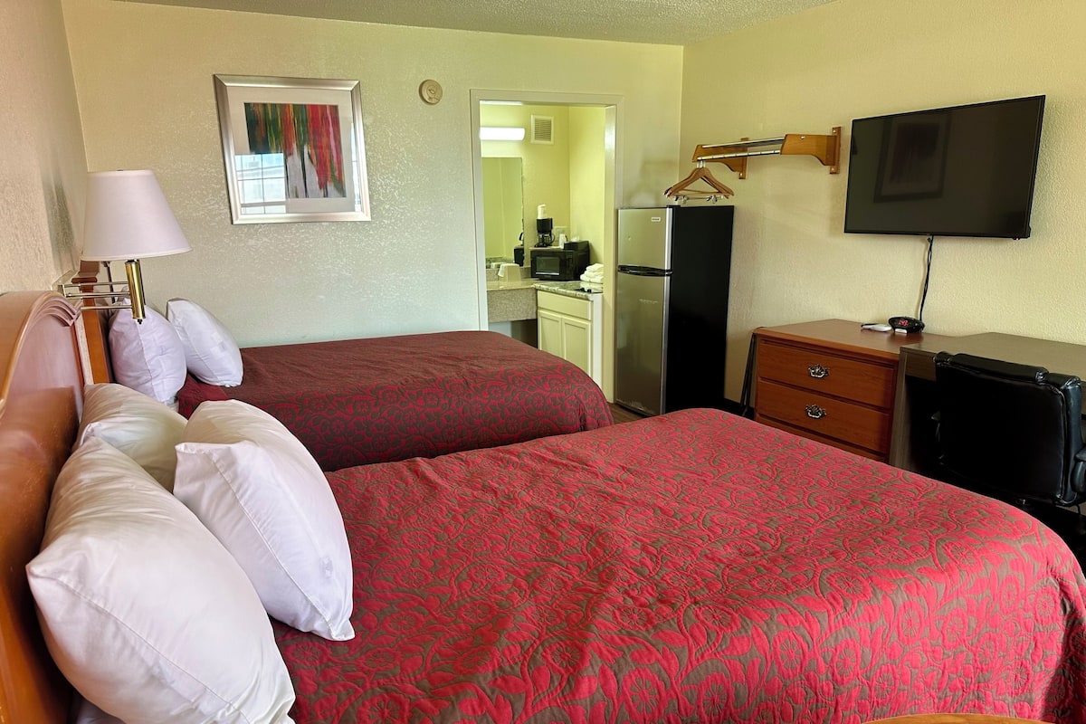 A hotel room with two beds, a desk, a TV, and a kitchenette.