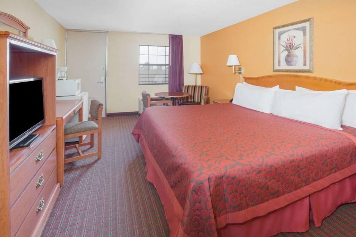 A hotel room with a large bed, a desk with a television, a chair, a small table, and a window with curtains.