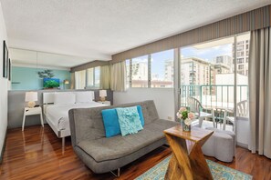 Ein Hotelzimmer mit Bett, einer Couch, einem kleinen Tisch mit Blumenvase, einem Balkon mit Geländer und Blick auf die Stadt.