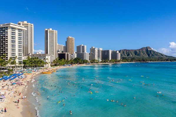 Ein Strand mit klarem türkisblauem Wasser, einem sandigen Ufer und einer Stadtansicht mit hohen Gebäuden im Hintergrund.