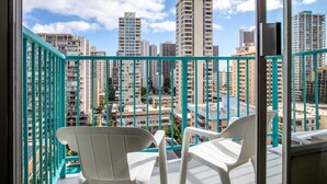 A balcony with white plastic chairs and a turquoise railing overlooking a cityscape with high-rise buildings.