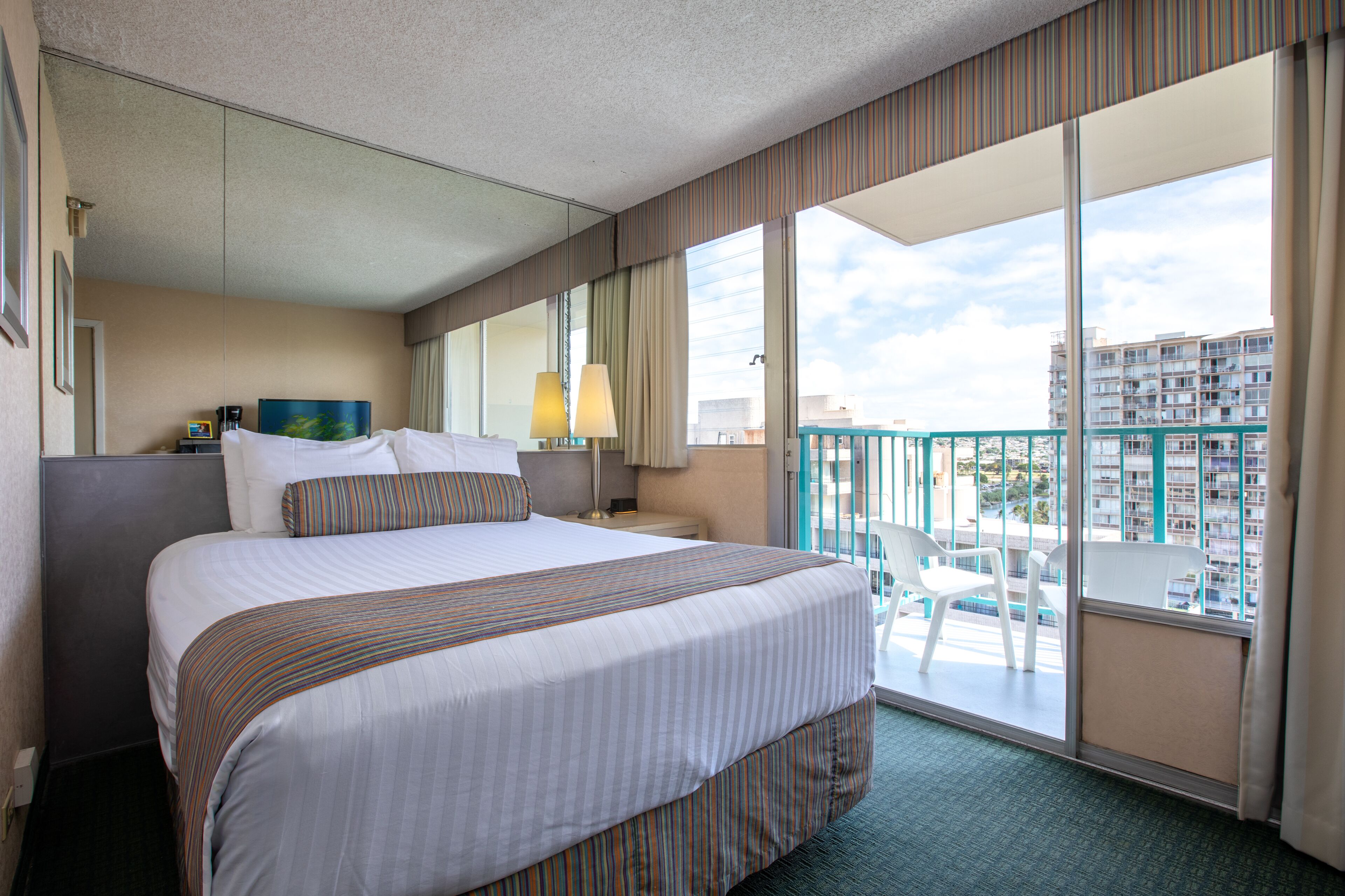 A hotel room with a large bed, a desk with a lamp, a view of the city through large windows, and a balcony with chairs.