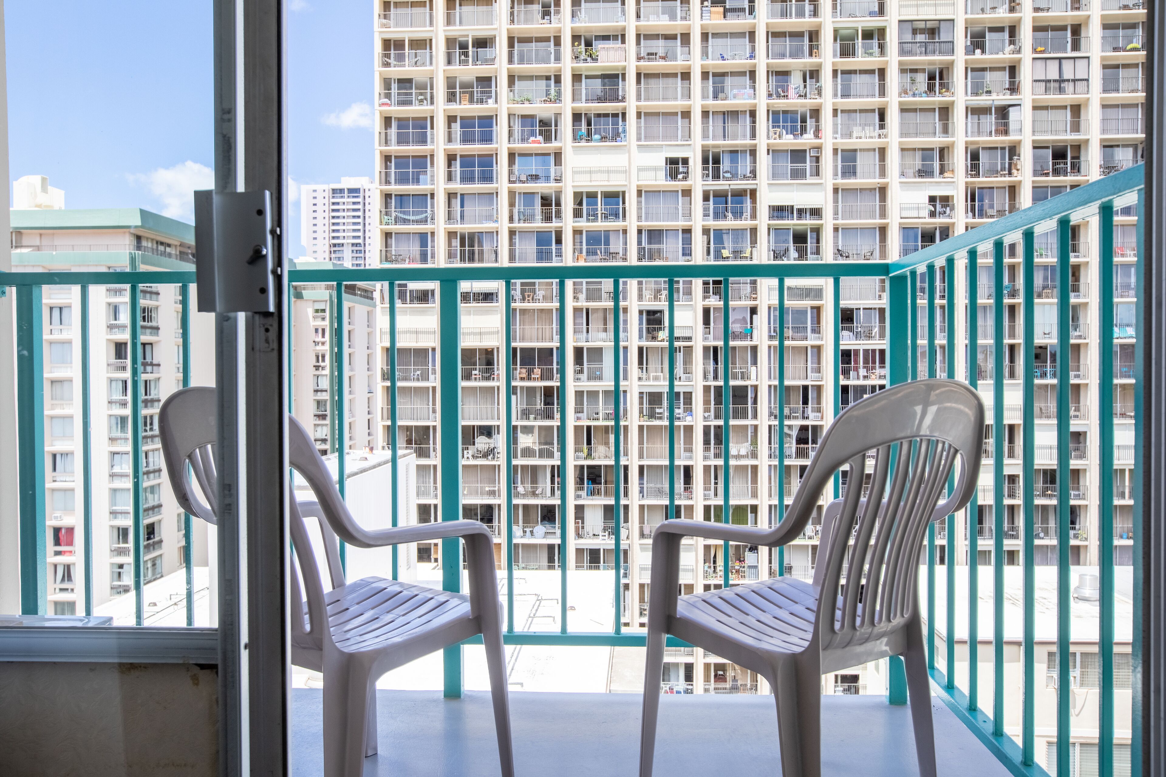 Een balkon met twee witte plastic stoelen, een metalen hekwerk en uitzicht op een hoog gebouw met meerdere balkons.