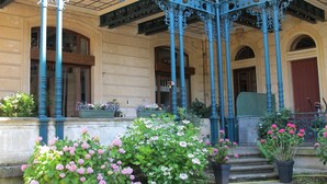 Un edificio con un portico coperto, caratterizzato da elaborate ringhiere e colonne in metallo blu, circondato da vasi con piante e fiori.