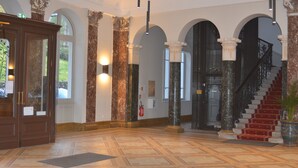 Un'imponente hall con colonne in marmo, una reception in legno e una scalinata con tappeto rosso.