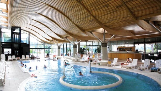 Un'ampia piscina interna con soffitto in legno curvo, circondata da poltrone relax e un'area bar.