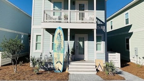 A two-story building with a balcony, a surfboard leaning against it, and a small front yard with plants. - Shore Do Love You: 5BR, Sleeps 12, Heated Private Pool & Optional Hot Tub! (Santa Rosa Beach)