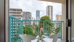 Een balkon met turquoise relingen, witte kunststof stoelen en uitzicht op hoge gebouwen en palmbomen.