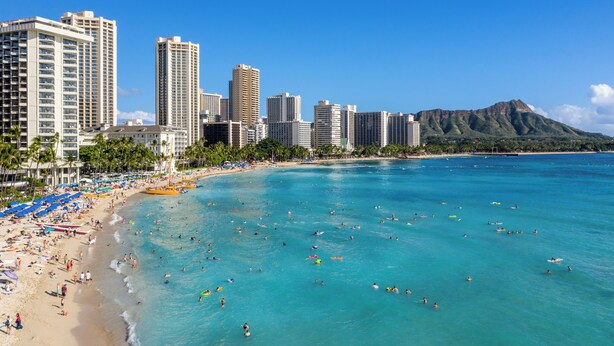 Una playa con aguas turquesas cristalinas, una orilla arenosa y un horizonte urbano con edificios altos al fondo.