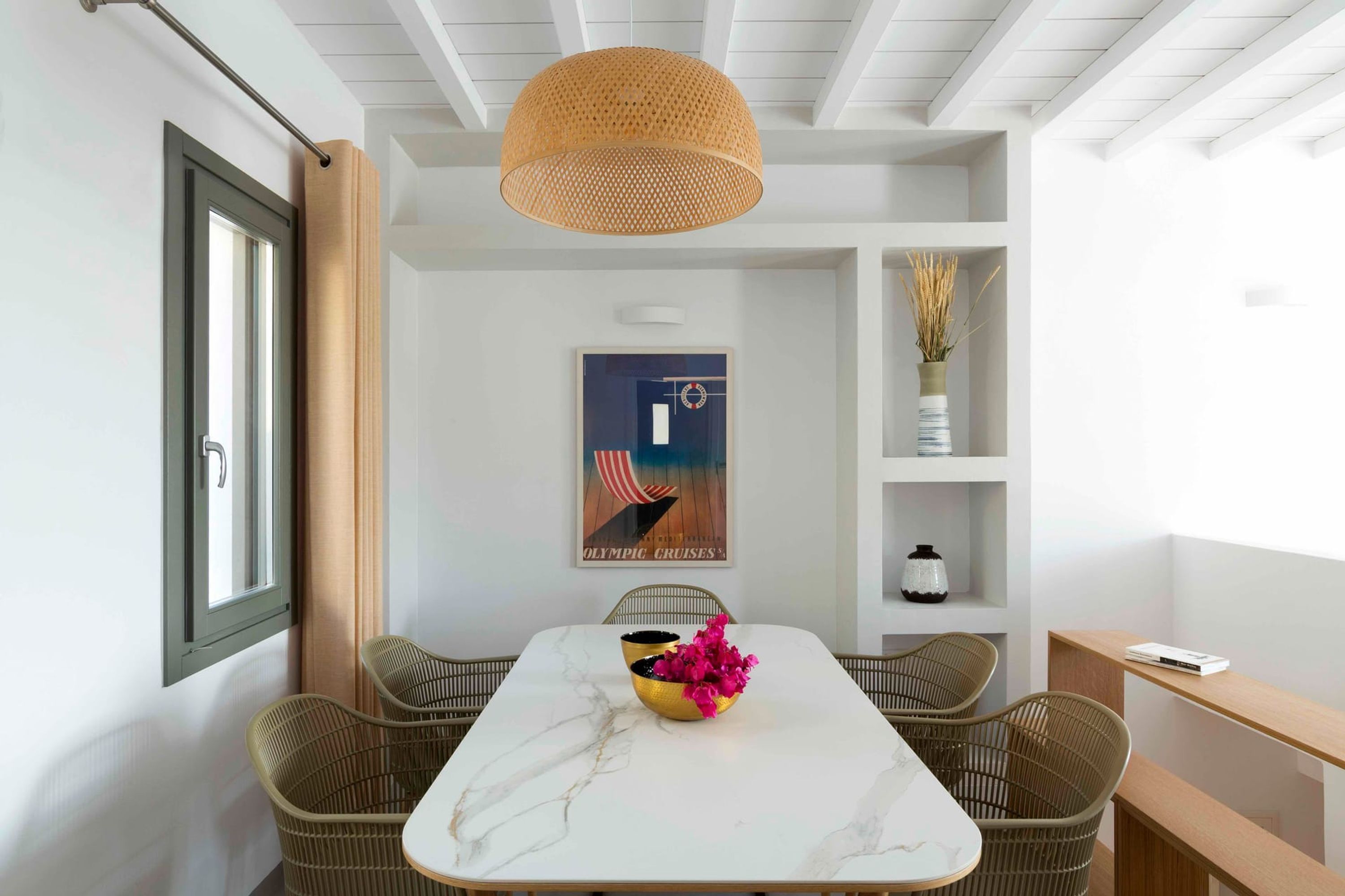 A dining area with a white marble table, wicker chairs, a hanging rattan light fixture, and a poster on the wall.