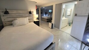 Une chambre d’hôtel moderne avec un grand lit, des tables de chevet, une commode, un miroir, une armoire et une salle de bain visible à travers une porte ouverte.