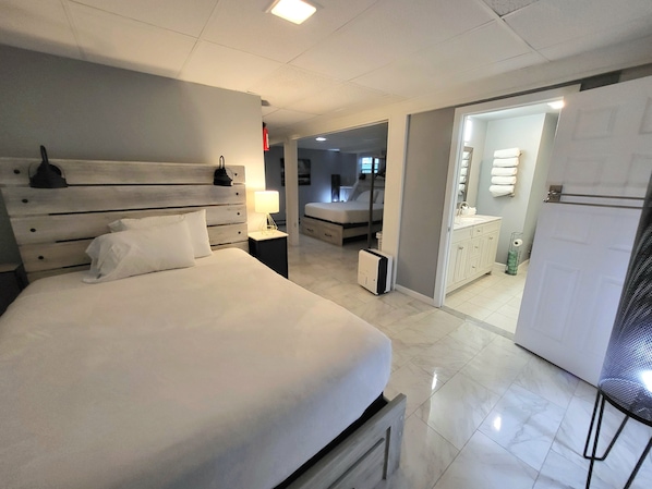 Et moderne hotelværelse med en stor seng, sengeborde, et natbord, et spejl, et garderobeskab og et badeværelse, der kan ses gennem en åben dør.