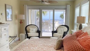Een slaapkamer met een bed, twee stoelen, een dressoir en uitzicht op de oceaan door grote ramen.
