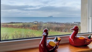 A view through a window of a coastal landscape with a distant island, a grassy area, and a body of water. - 4 bedroom accommodation in Lendalfoot, near Girvan (Lendalfoot, near Girvan)