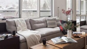 En moderne stue med en grå sofa, et træbord og et stort vindue med udsigt til et sneklædt landskab.