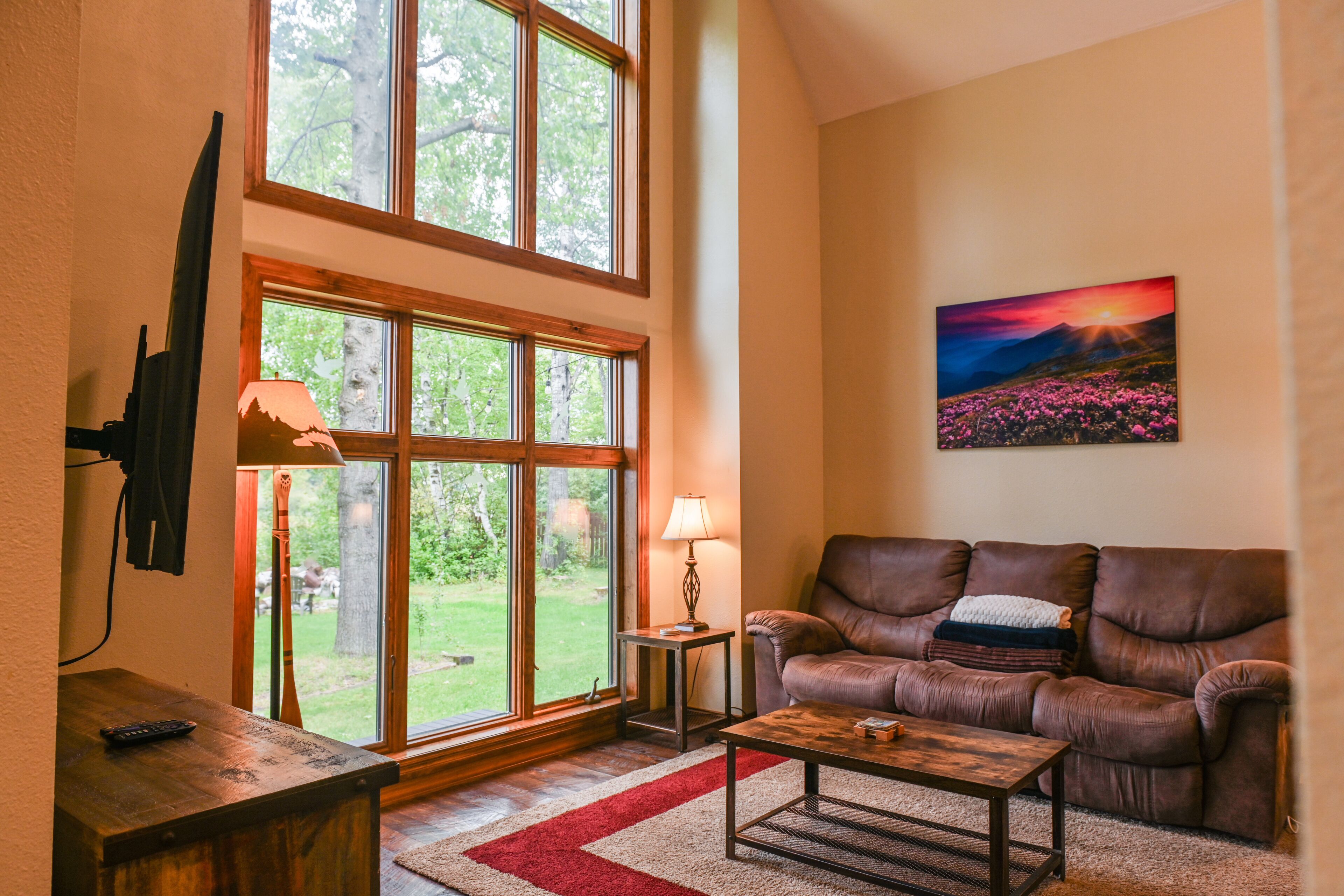 A cozy living room with a large window, a brown leather sofa, a coffee table, and a painting on the wall.