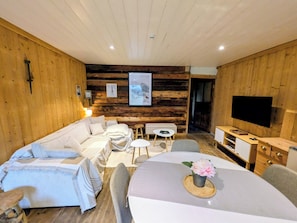 A cozy room with a wooden wall, a sofa set, a dining table with a flower centerpiece, and a flat-screen TV mounted on the wall.