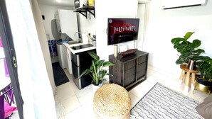A modern kitchen with a built-in oven, a microwave, and a sink. There is a flat-screen TV mounted on the wall displaying 'PARTY SONGS'. The room has a tiled floor and includes a woven pouf, a small wooden table, and a potted plant. - Ohhh-Our sweet little surprise #TinyPoolHouse #MiamiDesignDistrict (Miami)