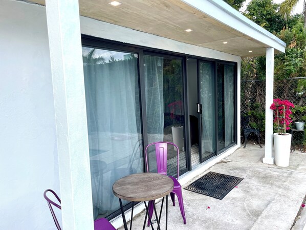 A modern building with large glass doors, a small patio with a round wooden table and purple chairs, and a potted plant with pink flowers.