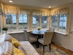 A dining area with a round table, patterned chairs, and a view of the outdoors through large windows. - Blue Water Shores Neighborhood walking distance to private inlet (Falmouth)