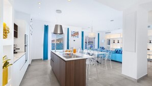 A modern kitchen with a central island, clear chairs, and a view of the sea.