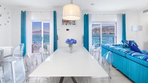 A modern room with a white table, clear chairs, a sofa with blue cushions, and a view of the sea.