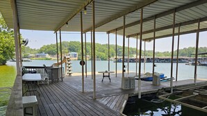 A wooden deck with a covered area, outdoor seating, and a view of a lake with boats and buildings in the background. - Lakefront Home with Private Dock, Water Slide, Boat Slip, and Hot tub (Lake Ozark)