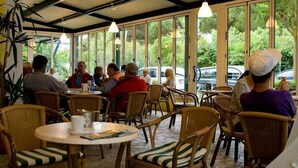 Un café al aire libre con mobiliario de mimbre y amplios ventanales que ofrecen vistas del entorno verde.