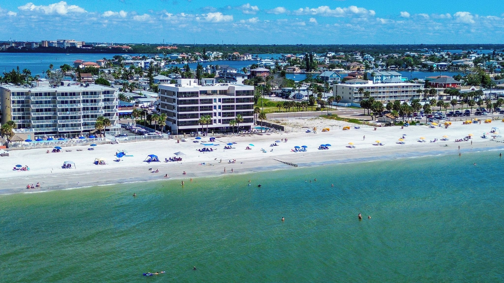 A coastal hotel with multiple floors, a beach in the foreground, and a marina in the background.