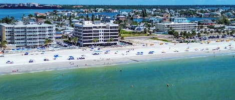 A coastal hotel with multiple floors, a beach in the foreground, and a marina in the background.