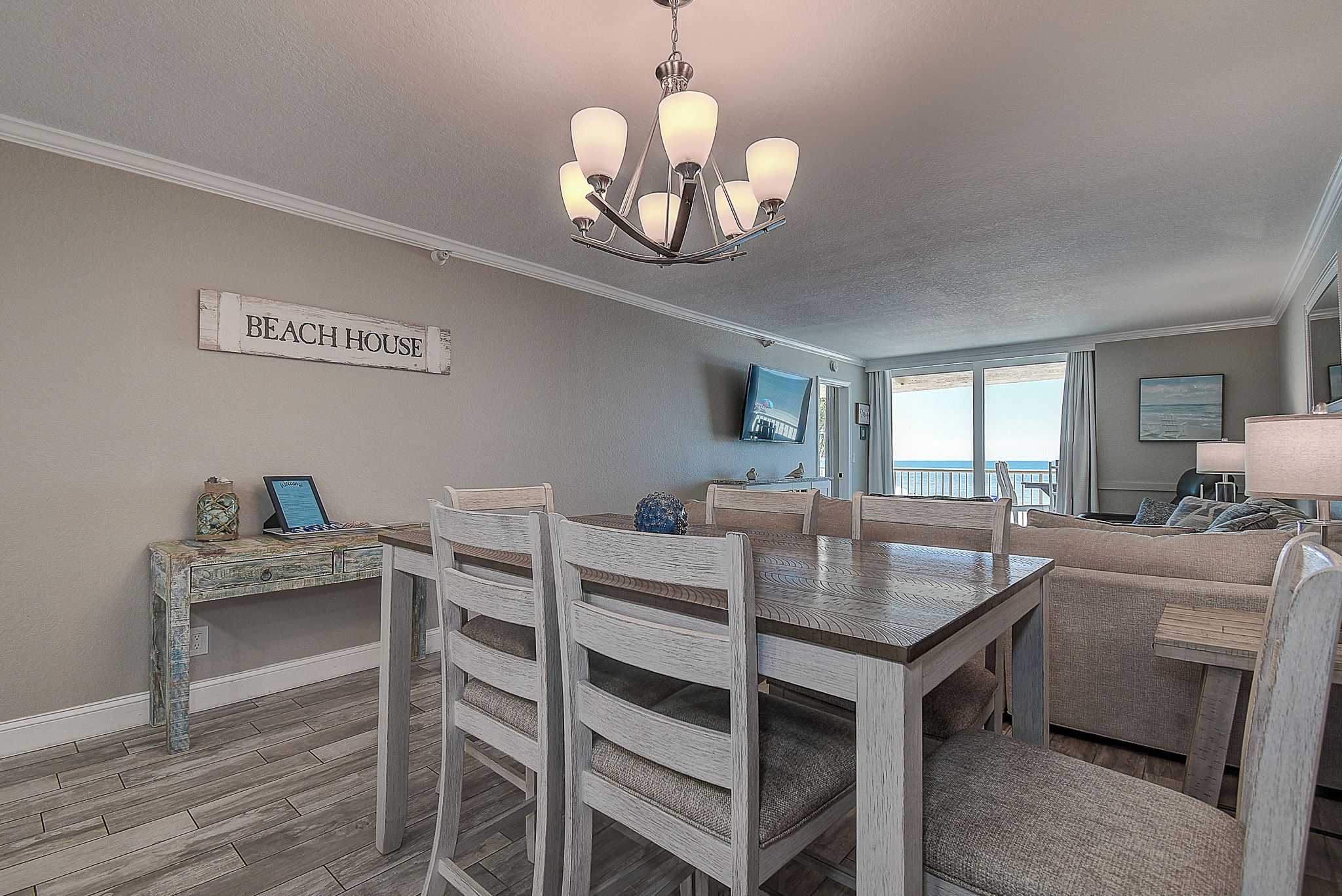 A dining area with a chandelier, a sign reading 'BEACH HOUSE', and a view of the beach through large windows.
