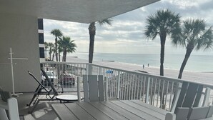 A covered outdoor seating area with a table and chairs, overlooking a beach with palm trees and the ocean.