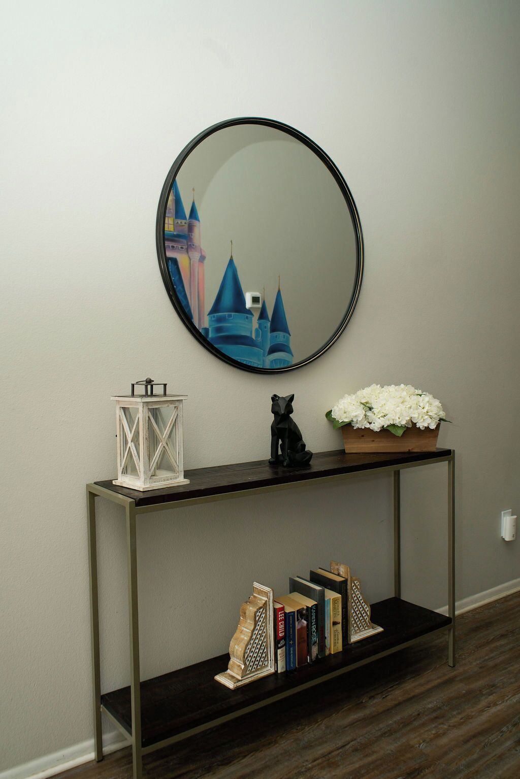 A hallway with a round mirror, a white lantern, a black cat statue, a flower arrangement, and a bookshelf with books.