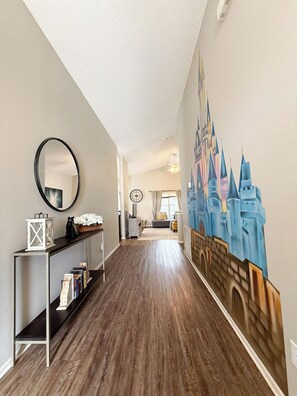 A hallway with a wooden floor, a mirror, a shelf with books, and a wall mural of a castle.