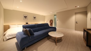 Une chambre d’hôtel moderne avec un canapé bleu, une table basse ronde en marbre et un lit recouvert de draps blancs.