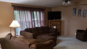 A living room with a brown sectional sofa, a lamp, a flat-screen TV on a wooden stand, and a ceiling fan. - Lodge at the Longbranch next to the Longbranch Saloon (AVA)