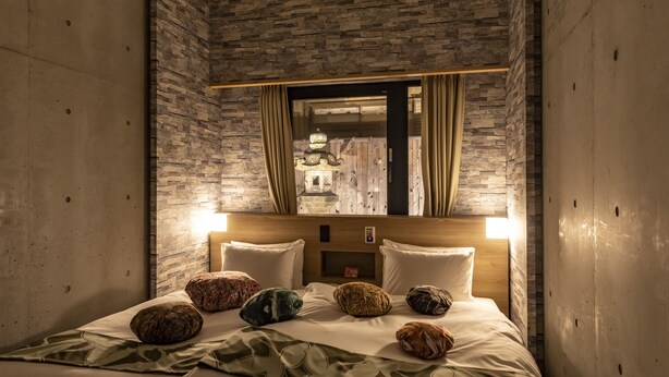 A hotel room with a stone wall, a bed with multiple pillows, a headboard with a mirror, and a window with curtains. - HALE Kyoto Tambaguchi (Kyoto)