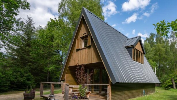 A wooden cabin with a sloped roof, a porch, and a barbecue grill in the foreground. - Rowan Cottage by Interhome (Drumnadrochit)